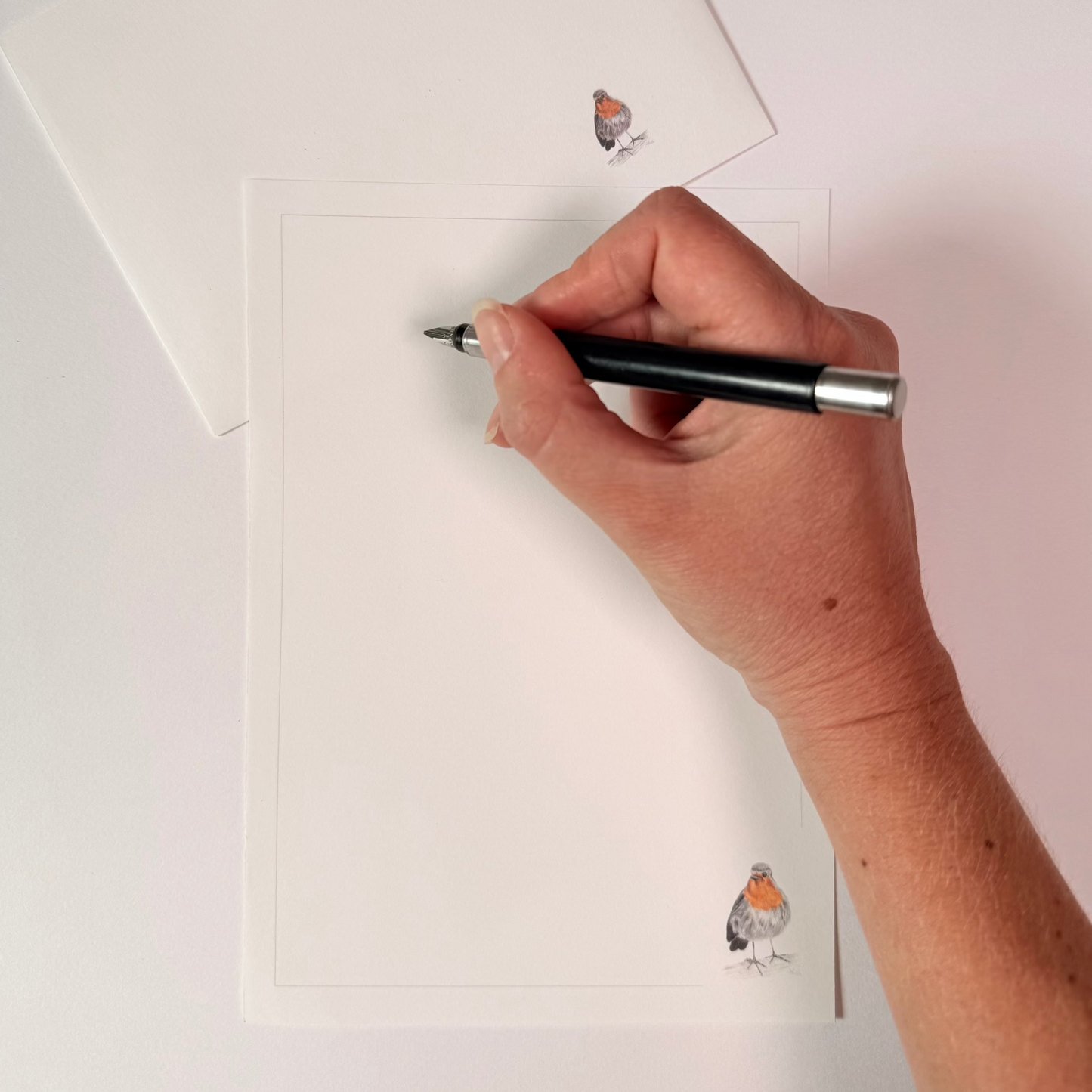 A hand holding a black and silver pen, writing on a white card with a delicate robin bird illustration in the bottom corner. A matching white envelope is partially visible in the top left corner.