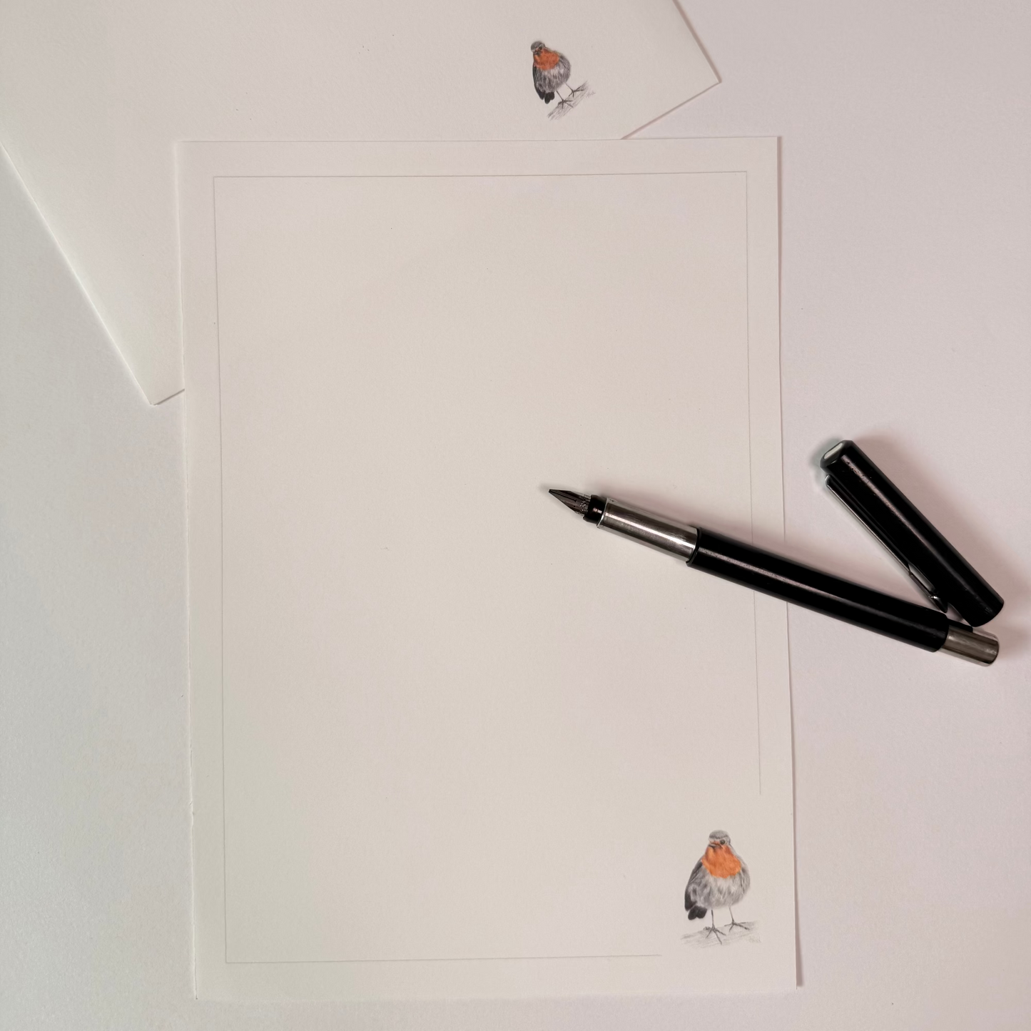 A white stationery set featuring a letterhead card and matching envelope with a small robin illustration in the bottom corner, styled with a black pen on a white background.