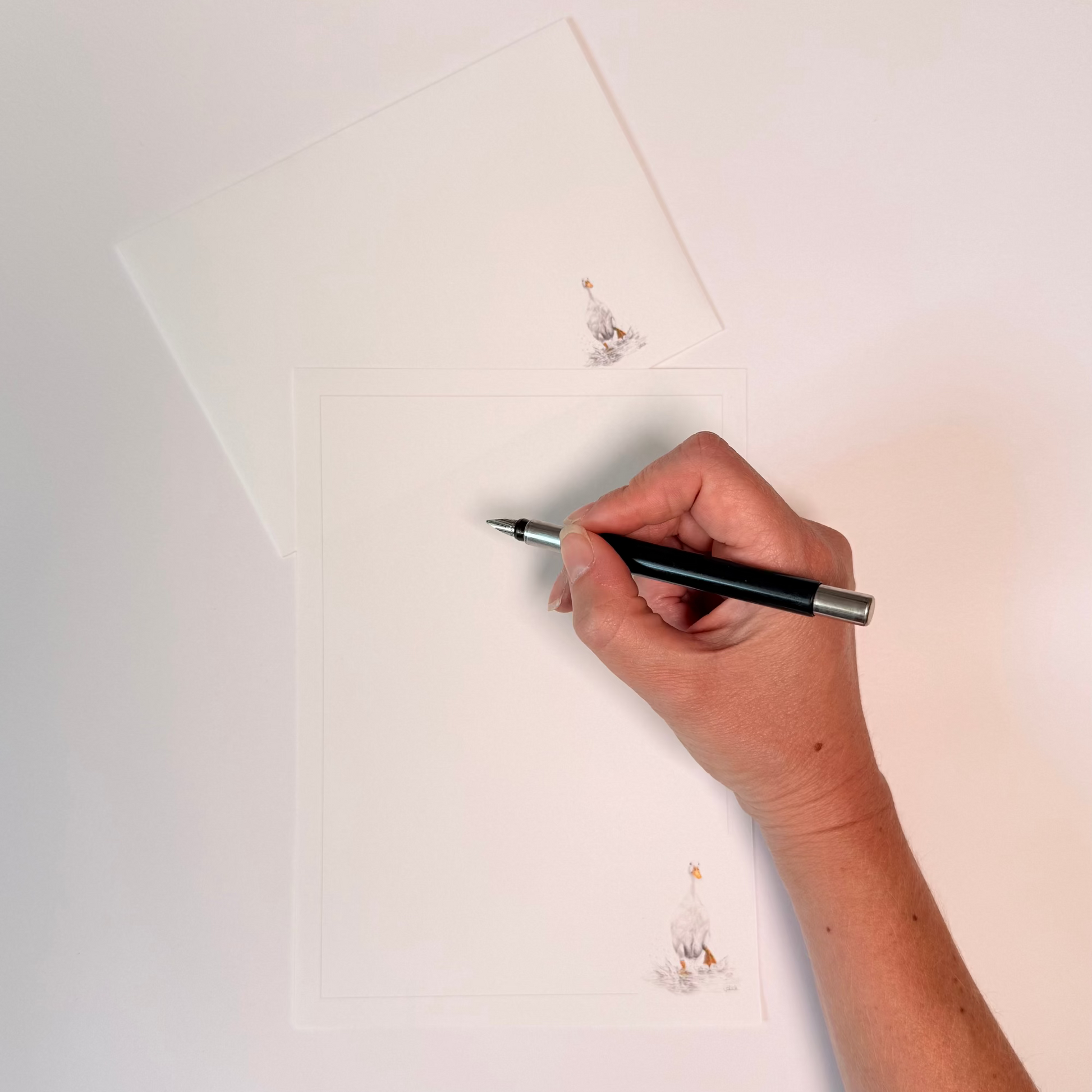 A hand holding a black and silver pen over white paper, with a matching white envelope or card placed above. Small printed illustrations appear near the top and bottom of the paper.