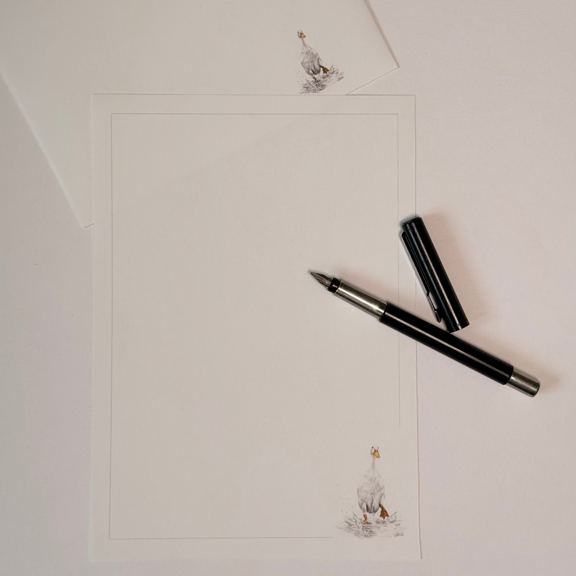 A flat-lay view of a white writing paper sheet with a thin border, decorated with small duck illustrations in the corners. A black and silver fountain pen and its cap are placed diagonally on the right side, and a matching paper envelope is visible in the top-left corner.